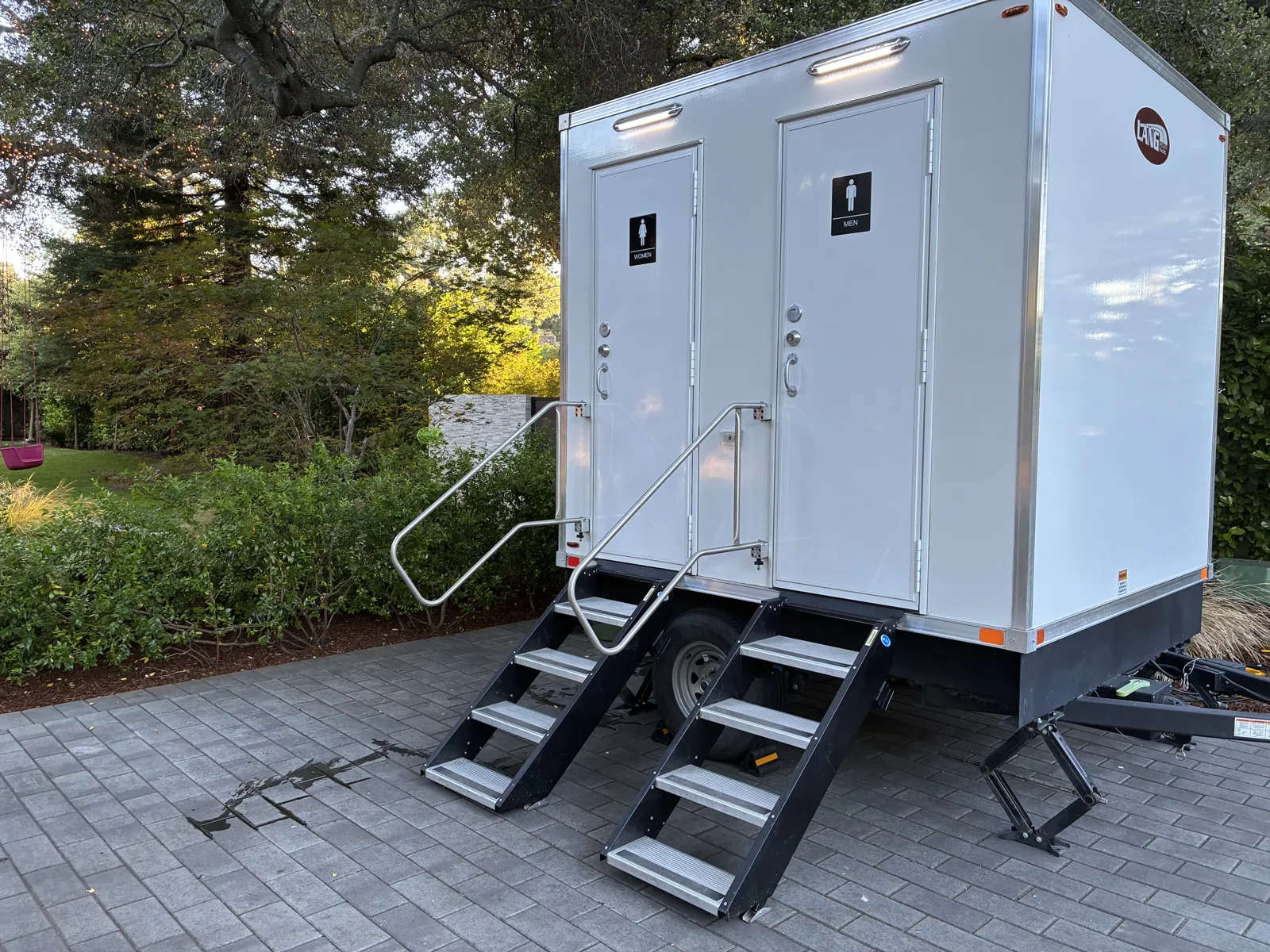 A white two station Pro Portable Restrooms luxury restroom trailer with steps in an estate garden