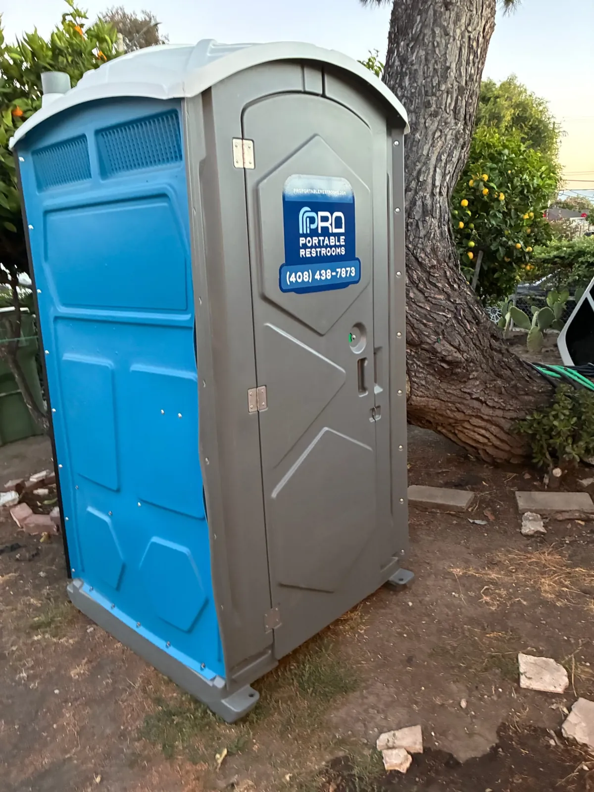 A clean blue Pro Portable Restrooms unit placed beside a tree at a Carmel outdoor event
